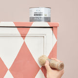 An Annie Sloan Clear Chalk Paint™ Wax tin placed on a Scandinavian Pink-and-Antoinette Pink diamond-patterned painted cabinet. A hand applies wax using a natural bristle brush, showcasing the sealing process.